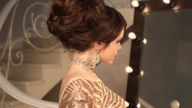 Happy young smiling girl with makeup and hairstyle, golden jewelry, red manicured nails. Attractive young brunette in sequin dress posing at camera in modern room.