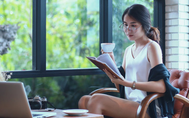 .Young pretty Asian girl sitting near opened window drinking coffee and reading book enjoys of rest,women working university project with generic design laptop. Analysis marketting plans,vintage color