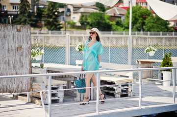 Naklejka premium Lovely girl wearing transparent dress, hat and sunglasses walking on the quay by the lake with her bag.