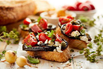 Toasts with grilled aubergine, cherry tomatoes, feta cheese, capers and fresh aromatic herbs, on a wooden table. Delicious Mediterranean appetizer