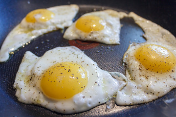 A close-up of four sunny-side up eggs frying in a pan with black pepper sprinkled on them