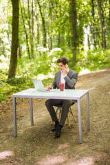 Fototapeta premium Businessman sitting at the office desk with laptop computer and cup of coffee talking mobile phone in green forest park. Business concept.
