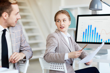 Pretty young white collar worker analyzing statistic data with her handsome colleague while sitting at desk in modern open plan office