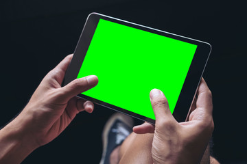 Mockup image of a man's hands holding black tablet pc with blank green screen on thigh in black background