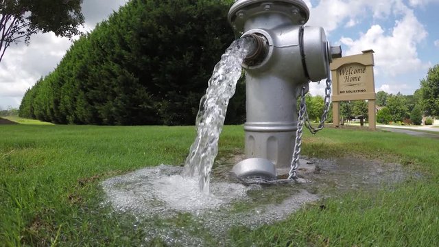 Opened silver fire hydrant with heavy flow clear water coming out puddling up around it and running down sidewalk near opening of community neighborhood.