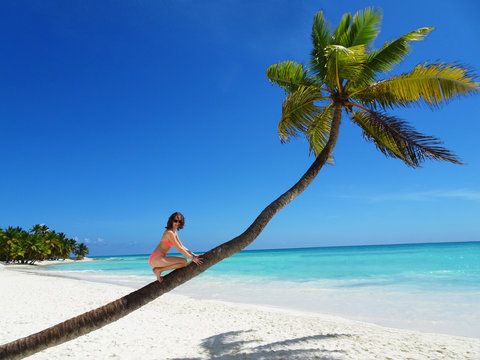 Junge Frau sitzt auf einer Palme am einsamen weißen Sandstrand auf der Insel Saona (Dominikanische Republik) und schaut lächelnd in die Kamera