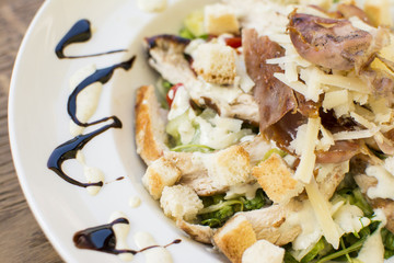 Healthy grilled chicken Caesar salad with cheese and croutons. Bacon and lettuce slices in a white plate on a wooden table.
