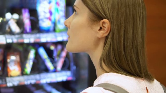 Beautiful hungry woman picking item out of vending machine in mall. Choosing unhealthy snacks being famished