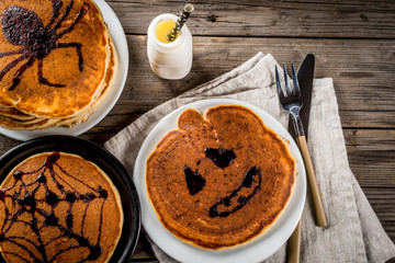 Ideas for breakfast are Halloween, food for children. Pumpkin pie pancakes decorated with chocolate syrup in a traditional style - spider web, spider, jack lantern. On wooden rustic table, copy space