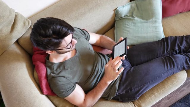 Handsome young man reading ebook on couch at home, wearing eyeglasses