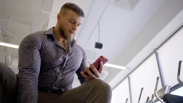 Business male bodybuilder trains biceps by lifting a dumbbell with one hand in business style clothes holding a mobile phone from the other hand dials the text message