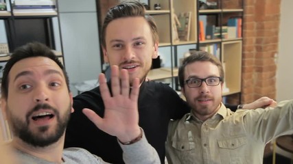 Young employees have fun in office of company indoors. They smile, wave their hands in front of camera and do selfi on occasion of successful completion of working week. Three men enjoy spending time