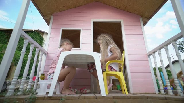 Backyard playground kids play house cubby. Children have fun together playing on the veranda of a wooden pink playhouse. Little girls drawing with color pencils outside.