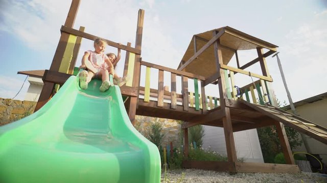 Happy children playing outside on playground slide in backyard. Two cute little girls have fun together sliding down in the garden. Low angle shot.