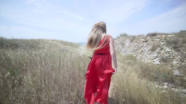 Young girl in red dress walking barefoot on the footpath towards the sea. Girl's long hair and bright scarlet gown blowing in the wind. Gimbal steadicam shot