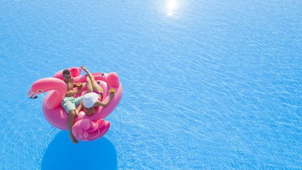 AERIAL CLOSE UP Beautiful young couple drinking cocktails, relaxing, lying on fun inflatable pink flamingo floatie. Sexy boyfriend and girlfriend sipping drinks, enjoying floating on sunny pool water
