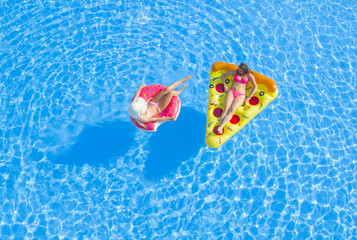 AERIAL TOP DOWN Relaxed girls in pink bikini swimsuits lying on fun inflatable pizza and flamingo floating pillows on water. Girlfriends on summer vacation enjoying colorful floats in swimming pool