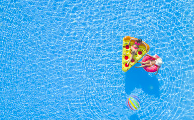 AERIAL TOP DOWN Relaxed girls in pink bikini swimsuits lying on fun inflatable pizza and flamingo floating loungers on water. Girlfriends on summer vacation enjoying colorful floaties in swimming pool