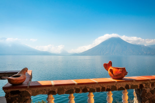 Hazy view of the blue waters of Lake Atitlan in the morning from a sunny terrace with some interesting ceramic decorations. In the background we can see the volcanoes San Pedro and Toliman.