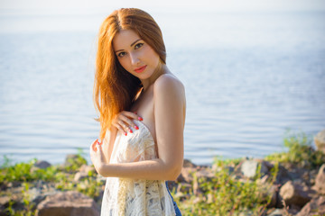 Fototapeta na wymiar Tender beautiful red hair lady at evening sunlight around rocks and sea water. Woman wearing in lace cardigan. Portrait in vintage style on vacation. Girl relax at warm day