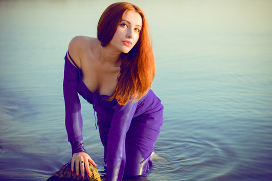 Gorgeous sexy young lady at evening sunlights around rocks and water. Woman wearing in purple long dress. Portrait in vintage style. Sweet woman rest on the sea