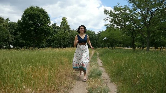 A splendid young woman in a folk dress with open shoulders and sandals runs along a country road and smiles happily in slow motion. She moves along a nice lawn with green trees on a sunny day