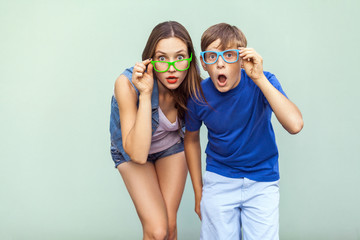 Eyewear concept. WOW faces. Young sister and brother with freckles on their faces, wearing trendy glasses, posing over light green background together. Looking at camera with surprised face.