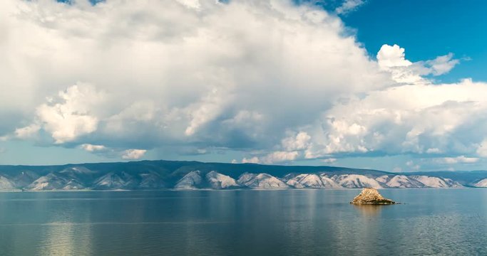 Timelapse. White clouds float above the smooth lake. A yellow rock above the calm water of the blue lake is a baikal. Rocks on the background. Fast moving clouds over the lake Baikal.