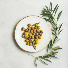 Pickled green Mediterranean olives in virgin olive oil on white ceramic plate and olive tree branch...