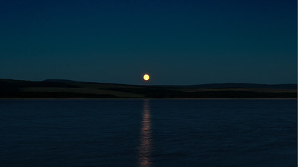 The moon rises from behind the hills above the surface of the lake leaving the lunar path