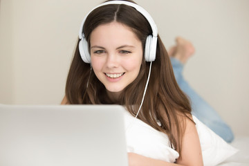 Smiling young woman wearing headphones lying on bed with laptop, teenage student studying online at home, listening to audio course, fast learning foreign language at home, head shot portrait