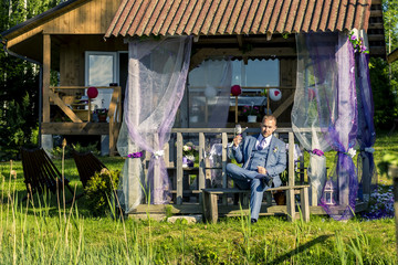 Portrait of young and attractive man in grey blue suit sitting next to the summerhouse and holding a glass of champagne.