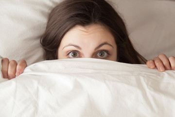 Funny surprised girl covering half of face with white blanket, young scared woman hiding and peeking from duvet, afraid of night monsters, feels embarrassed, wide awake, head shot close up, top view