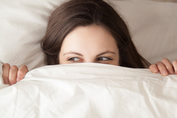 Playful young woman hiding face under blanket while lying in cozy bed on white pillow, pretty curious girl feeling shy peeking from duvet, covering with white sheet, head shot close up, top view