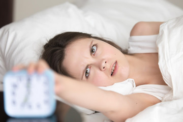 Fototapeta na wymiar Annoyed sleepy young woman reaching to ringing alarm clock to turn it off lying in bed in the morning, frustrated teenager woke early by annoying signal, waking up tired after bad sleep, head shot