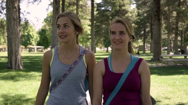 Young Women Walk Through Park With Yoga Mats Strapped To Their Backs, Woman On Right Tells A Surprising Story, They Laugh