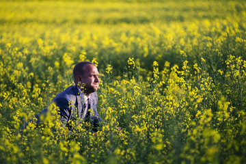 Fototapeta premium Young and attractive man in a suit sitting in yellow rape field and looking somewhere.