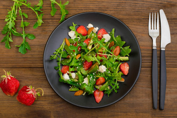 Salad with arugula, strawberries, cottage cheese, olive oil, on a black plate, wooden background. Close-up. Top view