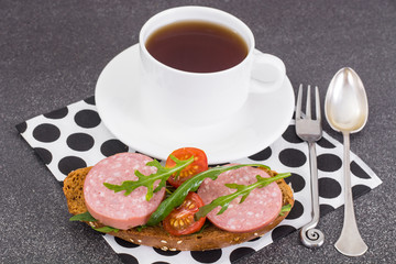 Tea in white cup and sandwich with sausage and tomatoes
