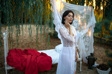 Young sexy woman stands near bed with bedding and baldachin on sand near tree. © Stanislav