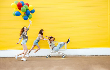 Smiling women friends have fun with shopping trolley and balloons.