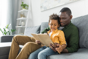 relaxed african-american father and son reading book together on sofa