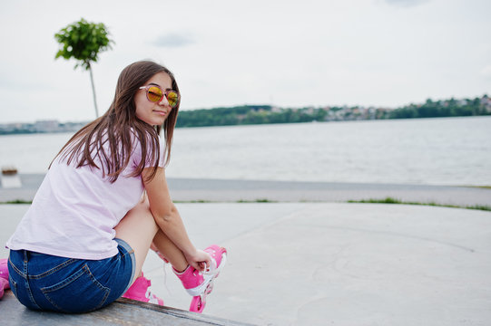 Portrait of a beautiful girl wearing sunglasses, t-shirt and shorts putting on rollerblades outdoor next to the lake.