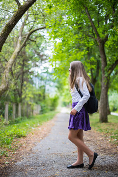 Pretty beautiful blonde child girl cheerful and happily walking across street back to school enjoying through alley in the nature with backpack, glasses, purple skirt and white shirt