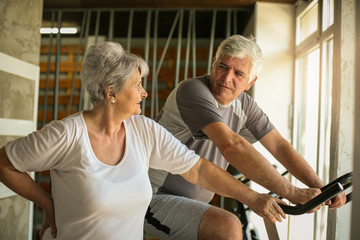 Fototapeta na wymiar Two senior people at the gym. Senior man sitting on the elliptical machine and having conversation with senior woman.