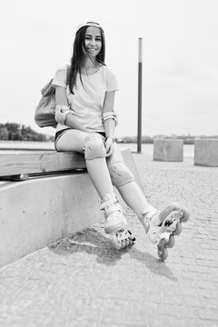 Fantastic young woman in casual clothing and cap sitting on the bench in the skatepark with rollerblades on. Black and white photo.