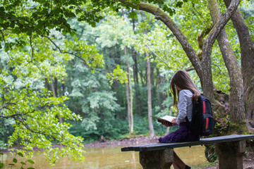 Pretty little blonde schoolgirl with backpack sitting on the bench in the nature park near lake and reading a book getting ready to go back to school in the nature studying for exam