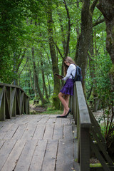 Pretty little blonde schoolgirl with backpack standing on a bridge in the nature and reading a book getting ready to go back to school in the nature studying for exam