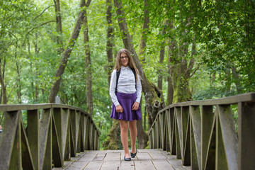 Beautiful pretty blonde school girl child smiling with glasses, white shirt, purple skirt and backpack taking a walk on the bridge in nature back to school autumn