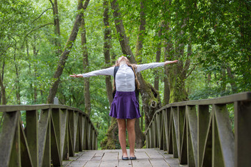 Beautiful pretty blonde school girl child smiling with glasses, white shirt, purple skirt and backpack standing on the bridge in nature cheerful arms widespread back to school autumn